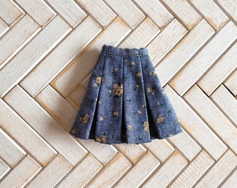 May include: A blue pleated skirt featuring a small, repeating floral pattern in shades of beige and light green. The skirt has a structured waistband and a flared silhouette, with visible vertical pleats running from the waist to the hemline.