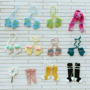 Pode incluir: Uma coleção de acessórios de inverno feitos à mão, incluindo luvas, um cachecol, meias e uma flor. As luvas vêm em várias cores como azul, verde, rosa e amarelo. As meias são amarelas, rosas e pretas. Os itens são dispostos em uma superfície branca texturizada.