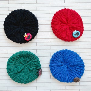 May include: Four knitted beanies in different colors: black, red, green, and blue. Each beanie has a different character button attached: Elmo, Cookie Monster, a green monster, and a brown button.