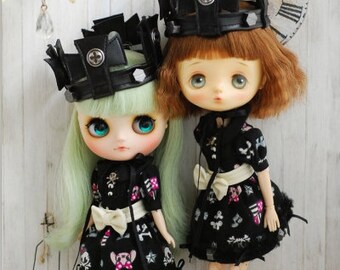 May include: Two dolls with large eyes and black crowns. One doll has mint green hair, the other has auburn hair. Both wear black dresses with white graphic patterns, white waist bows, and black shoes. The doll on the right wears black and white patterned socks.