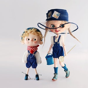 Op de afbeelding: Twee poppen in spijkerbroek-overalls en hoeden. E&eacute;n pop draagt een rood-wit gestreepte sjaal en een blauwe gebreide muts. De andere pop heeft lange blonde vlechten, een blauwe emmer en een visnet. De poppen staan op een witte achtergrond.