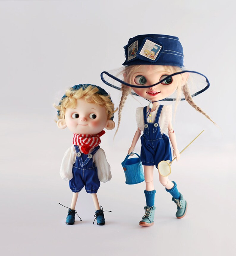Op de afbeelding: Twee poppen in spijkerbroek-overalls en hoeden. E&eacute;n pop draagt een rood-wit gestreepte sjaal en een blauwe gebreide muts. De andere pop heeft lange blonde vlechten, een blauwe emmer en een visnet. De poppen staan op een witte achtergrond.