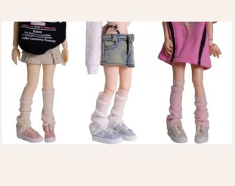 Könnte beinhalten: Drei Puppen in verschiedenen Outfits und weißen Kniestrümpfen mit Spitzenbesatz. Die Puppen tragen Jeans-, Khaki- und rosa Röcke. Die Puppen tragen weiße Turnschuhe.