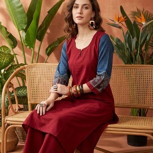 May include: A woman wearing a burgundy kurta and matching palazzo pants. The kurta has blue sleeves with white polka dots and patterned cuffs. She is seated on a wicker chair, with plants in the background.