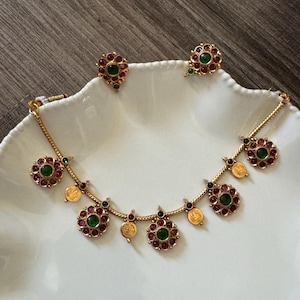 May include: A gold-toned necklace and matching stud earrings set. The necklace features a chain with alternating circular floral pendants containing green and red stones, interspersed with small gold coin charms. The earrings match the floral design.