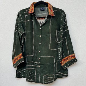 May include: A dark green button-down shirt featuring a white dotted geometric grid pattern. The collar and sleeve cuffs are accented with a contrasting orange and red floral border. The shirt includes a front button placket and a label reading FLO.