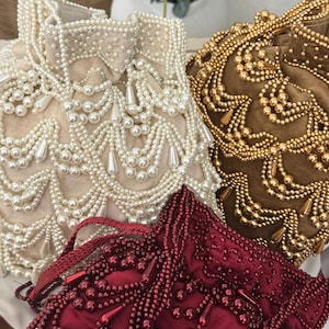 May include: Close-up of three beaded bags in cream, gold, and burgundy. The cream bag features cascading pearl-like beads. The gold bag has an intricate pattern of gold beads. The burgundy bag is adorned with red beads.
