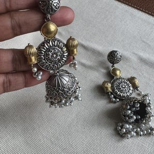 May include: A pair of silver and gold-toned earrings with intricate floral and geometric designs. The earrings feature a combination of textures, including a domed bell shape and small dangling pearls. The earrings are displayed against a neutral background.