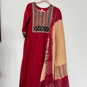 May include: A red tunic dress with a decorative neckline and three-quarter sleeves, paired with a beige and red patterned scarf. The neckline features vertical stripes and embroidered details. The scarf has a mix of stripes and paisley patterns.