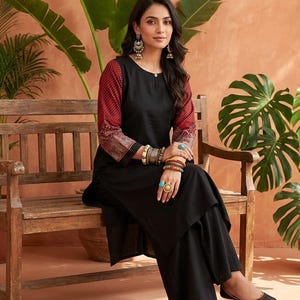 May include: A woman wearing a black kurta and palazzo pants, seated on a wooden bench. The kurta features red patterned sleeves. She is wearing silver earrings, bracelets, and rings. The background includes large green plants and a peach-colored wall.