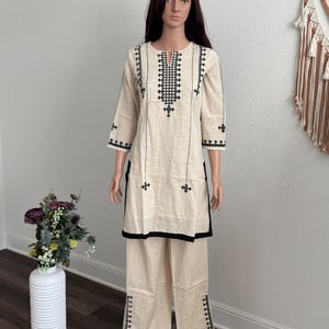 May include: Cream-colored two-piece outfit with black embroidered details. The top has a square neckline and three-quarter sleeves. The pants are wide-legged. The outfit is displayed on a mannequin.