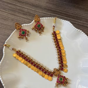 May include: A gold-toned necklace and matching earring set featuring square red stones, small circular gold coins, and tiny white beads. The necklace includes a central pendant with a green stone, while the earrings feature a similar design.