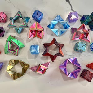 May include: A collection of colorful, geometric puzzle toys in various shapes, including cubes, stars, and multi-sided figures. The toys feature iridescent and metallic finishes in shades of pink, blue, green, and red. These brain teaser toys are designed for fun and mental stimulation.