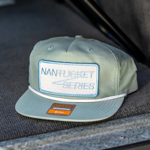 May include: A light green Nantucket Series baseball cap with a white rope detail and a rectangular patch. The patch features the words "NANTUCKET SERIES" in white and blue. The cap has a curved brim and a Richardson tag.