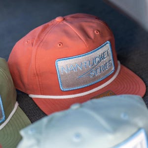 Puede incluir: Gorra de béisbol de color coral con un detalle de cuerda blanca y un parche rectangular. El parche presenta el texto "Nantucket Series" en bordado azul claro. La gorra tiene una visera curva y un botón en la parte superior.
