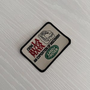 May include: A beige patch with black trim featuring the text "1994 LA RUTA MAYA AN EXPEDITION OF DISCOVERY" in red and black. A Land Rover logo is on the bottom right. The patch has a diamond shape.