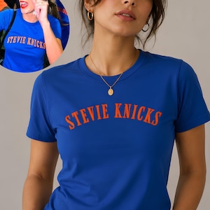 Puede incluir: Una camiseta de manga corta de color azul real con el texto STEVIE KNICKS impreso en una fuente naranja arqueada y llamativa en el pecho. El diseño es consistente con la camiseta que se muestra en la imagen circular, que exhibe la misma prenda.