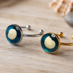 Puede incluir: Dos anillos abiertos con engastes circulares, incrustaciones de resina azul profundo y una pequeña concha espiral de color crema incrustada en el centro. Un anillo tiene una banda de metal plateado y el otro una banda de metal dorado.