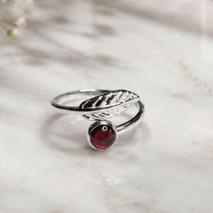 May include: A silver ring featuring an open band design with a polished metal leaf motif on one end and a single, round, deep red garnet gemstone set in a smooth silver bezel on the other end, creating a bypass style ring with a reflective finish.