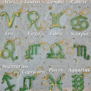 May include: A collection of twelve zodiac sign keychains. Each keychain features a green and gold glitter design with a gold-colored keyring. The signs are labeled: Aries, Taurus, Gemini, Cancer, Leo, Virgo, Libra, Scorpio, Sagittarius, Capricorn, Pisces, and Aquarius.