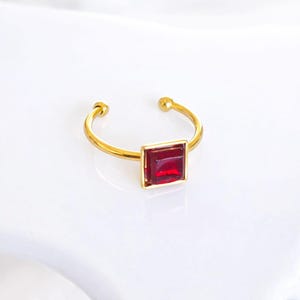 May include: A gold ring featuring a square, deep red gemstone. The ring has a simple, open design with small, round details at the ends. The gemstone is set within a gold frame, creating a striking contrast.