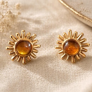 May include: A pair of gold-toned stud earrings shaped like suns, each featuring a central round amber-coloured stone surrounded by radiating metal rays. The earrings have a polished metallic finish and a circular design that mimics a sunburst.