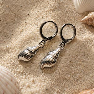 May include: A pair of silver-toned hoop earrings featuring dangling seashell charms. Each earring consists of a circular hinged hoop connected to a small, detailed metallic shell pendant with a polished finish and visible ridges along the surface.