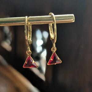 Könnte beinhalten: Ein Paar goldfarbene Klappcreolen mit dreieckigen, tiefroten Edelsteinen. Die Edelsteine sind in einen goldfarbenen Rahmen gefasst, was einen auffälligen Kontrast erzeugt. Die Ohrringe werden auf einem goldfarbenen Stab präsentiert.