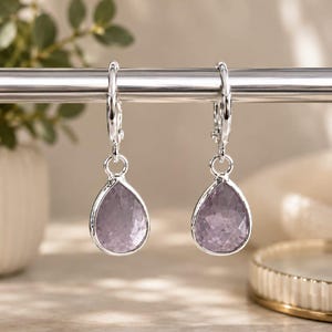 May include: Two silver hoop earrings, each with a teardrop-shaped, faceted, lavender gemstone. The earrings are suspended from a silver bar, with a blurred background including a plant and a decorative dish.