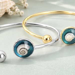 May include: Two open cuff bracelets, one in silver-tone metal and one in gold-tone metal. Each bracelet features a circular charm containing a small, spiraled seashell encased in a deep teal, translucent resin dome and a smooth metal ball end.