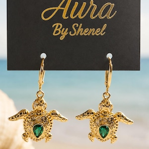 Peut inclure: Une paire de boucles d'oreilles pendantes dorées en forme de tortue de mer, ornées d'une pierre verte en forme de poire et de petits accents clairs. Elles sont fixées sur un support noir avec le texte doré Aura By Shenel imprimé dessus.