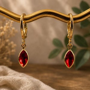May include: A pair of gold-toned hoop earrings featuring dangling marquise-cut red gemstones. Each earring consists of a polished gold circular hoop with a hinged closure, suspending a single faceted, deep red stone set in a gold metal frame.