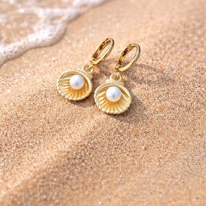 May include: A pair of gold-coloured seashell earrings, each with a white pearl centre, set on a sandy beach. The earrings feature a detailed, textured design and a hoop fastening. The background shows the sea and sand.