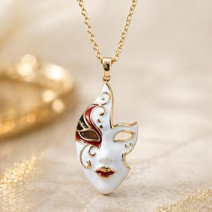 Op de afbeelding: Goudkleurige ketting met een witte maskerhanger met rode, zwarte en gouden accenten. Het maskerontwerp omvat een gestileerd oog, lippen en decoratieve krullen. De ketting is een fijne schakelstijl.