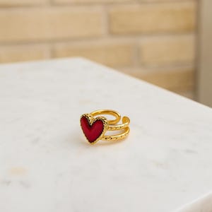 May include: A gold-coloured ring featuring a red heart-shaped centre. The ring has a double-band design with a textured pattern. The heart is a vibrant red colour, and the ring appears to be made of metal.