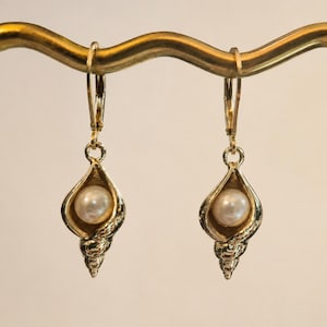 May include: A pair of gold-toned drop earrings featuring a textured seashell charm with a central white pearl. Each earring is suspended from a lever-back closure, showcasing a detailed spiral shell design that cradles the round pearl securely.