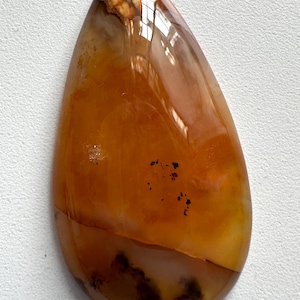 May include: Polished, teardrop-shaped gemstone with a warm color palette. The stone features a blend of orange, brown, and cream hues, with darker inclusions near the base. The surface is glossy, reflecting light, and the overall shape is smooth and organic.