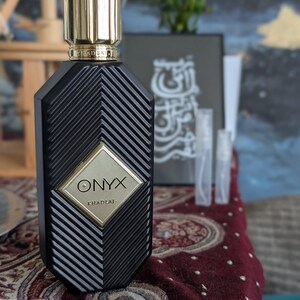 Peut inclure: Un flacon de parfum noir avec un bouchon dor&eacute; et le mot "ONYX" en lettres dor&eacute;es. Le flacon pr&eacute;sente un motif g&eacute;om&eacute;trique et repose sur une surface &agrave; motifs. L'arri&egrave;re-plan comprend une &oelig;uvre d'art encadr&eacute;e et de petits flacons d'&eacute;chantillons.