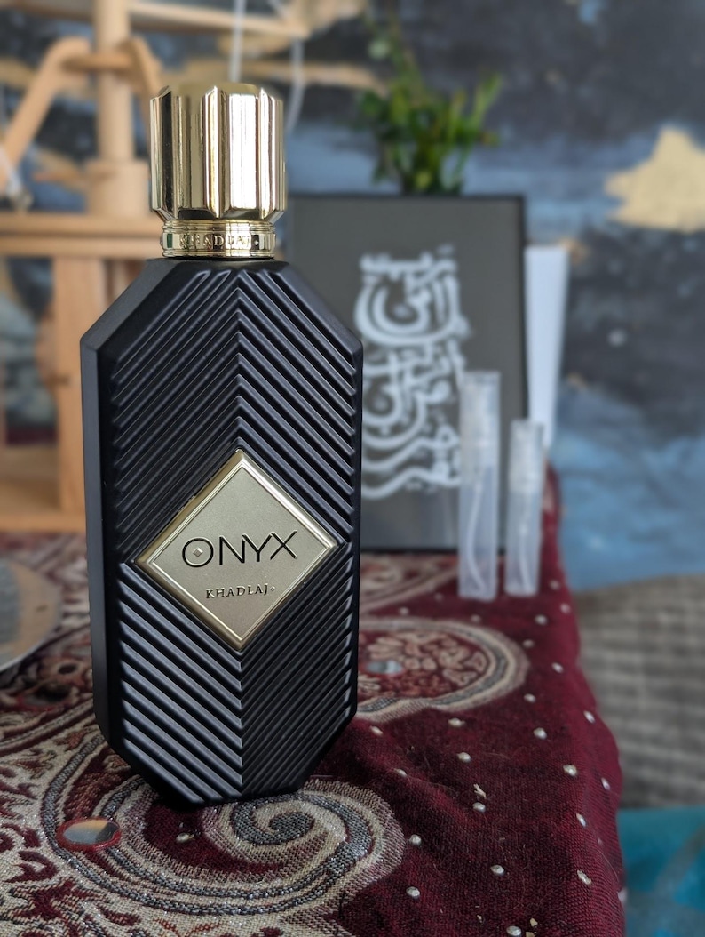 Peut inclure: Un flacon de parfum noir avec un bouchon dor&eacute; et le mot "ONYX" en lettres dor&eacute;es. Le flacon pr&eacute;sente un motif g&eacute;om&eacute;trique et repose sur une surface &agrave; motifs. L'arri&egrave;re-plan comprend une &oelig;uvre d'art encadr&eacute;e et de petits flacons d'&eacute;chantillons.