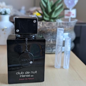 May include: A black rectangular perfume bottle with a silver cap and the text "Club de Nuit Intense Man" in white. The bottle has a silver chain and a round emblem with the brand name. Two clear spray bottles are next to it.