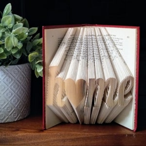 May include: A red hardcover book with the word "home" folded into the pages. The book is open and the pages are folded to create the word. The book is sitting on a wooden surface.
