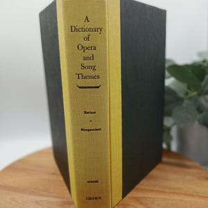 May include: A hardcover book titled "A Dictionary of Opera and Song Themes" by Barlow and Morgenstern. The spine is yellow with black text, and the covers are dark gray. The book is on a wooden surface.