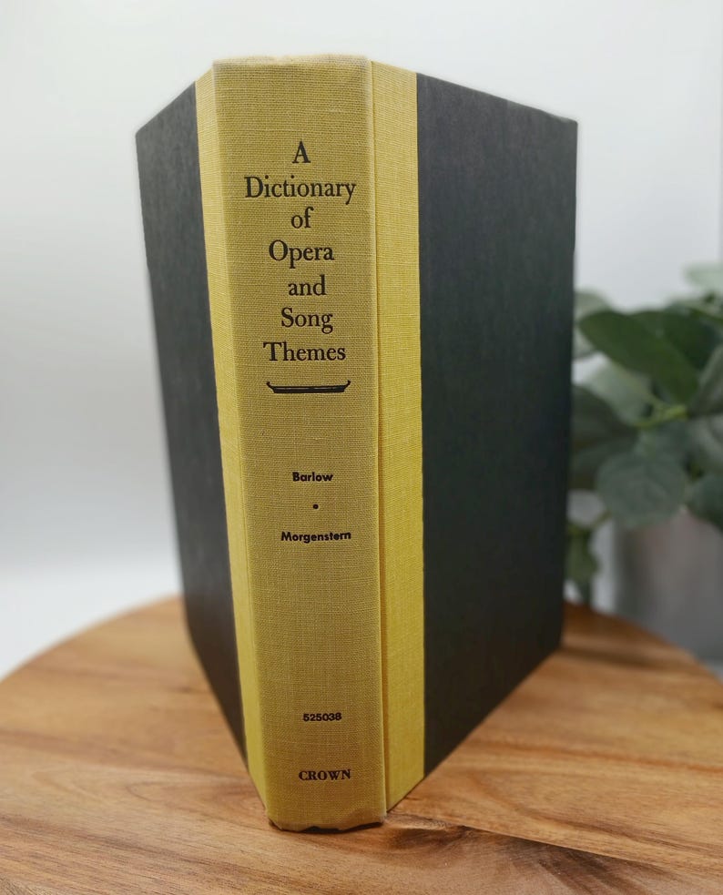 May include: A hardcover book titled "A Dictionary of Opera and Song Themes" by Barlow and Morgenstern. The spine is yellow with black text, and the covers are dark gray. The book is on a wooden surface.