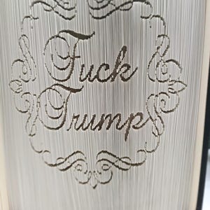 May include: A book with pages intricately cut to display the words "Fuck Trump" within a decorative frame. The text is in a serif font, and the book's pages are visible.