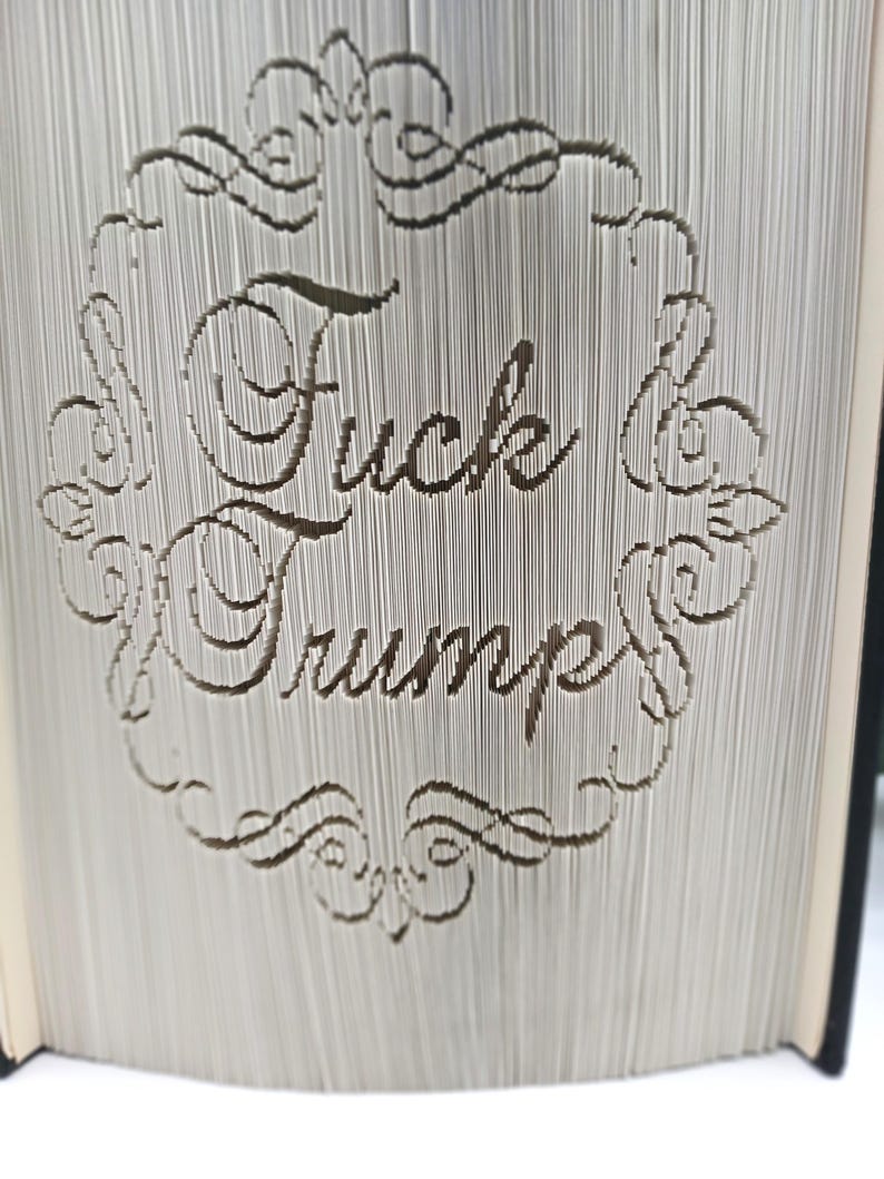 May include: A book with pages intricately cut to display the words "Fuck Trump" within a decorative frame. The text is in a serif font, and the book's pages are visible.