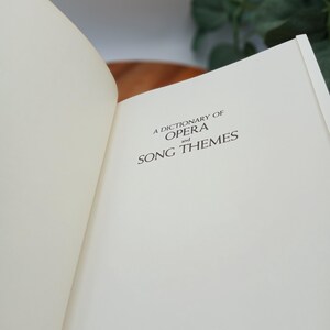 May include: Open book with the title "A Dictionary of Opera and Song Themes" printed in black on an off-white page. The book is open to a double-page spread, with the left page mostly blank. The background is blurred.