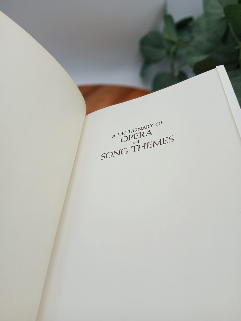 May include: Open book with the title "A Dictionary of Opera and Song Themes" printed in black on an off-white page. The book is open to a double-page spread, with the left page mostly blank. The background is blurred.