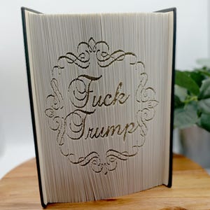 May include: A book with pages carved to display the words "Fuck Trump" within an ornate frame. The book's pages are light-colored, and the text is carved in a decorative font. The book is open, revealing the carved design.