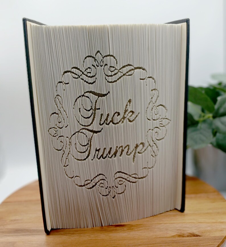 May include: A book with pages carved to display the words "Fuck Trump" within an ornate frame. The book's pages are light-colored, and the text is carved in a decorative font. The book is open, revealing the carved design.