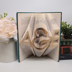 May include: A book art sculpture featuring the letters A, &, and E created by precisely folding the individual pages of a hardcover book. The cream-colored paper pages are fanned out to form the three-dimensional characters in the center.